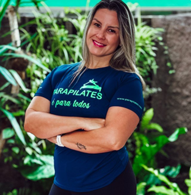 Vânia Cruz, de braços cruzados e camiseta azul-marinho da Parapilates, em um jardim.